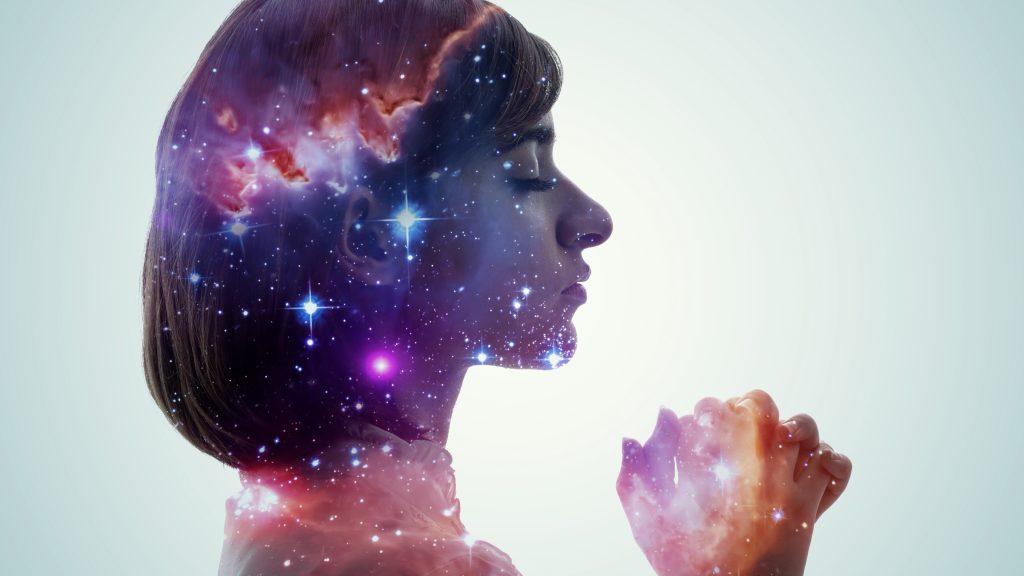 A woman in profile with a cosmic galaxy overlay, eyes closed, hands clasped in prayer against a light background.