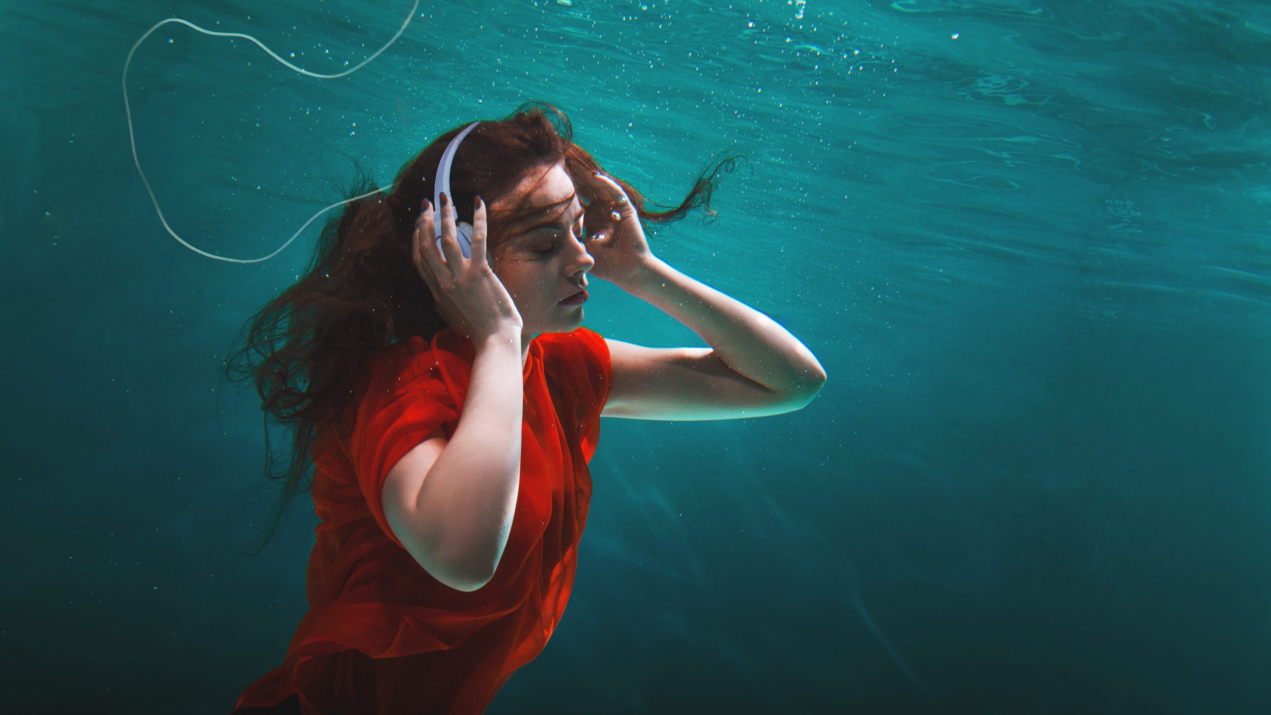 A woman in a red dress submerged underwater, wearing headphones, with flowing hair and a serene expression.