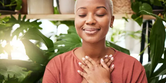 A woman with short blonde hair smiles peacefully, hands over her heart, surrounded by lush green plants.