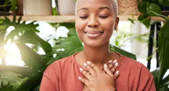 A woman with short blonde hair smiles peacefully, hands over her heart, surrounded by lush green plants.
