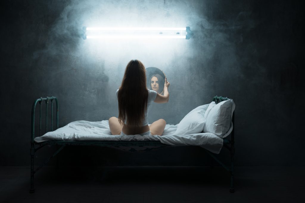 A woman sits on a bed, gazing into a mirror under a bright light, surrounded by a misty atmosphere.