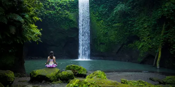 A woman sits on mossy rocks by a serene waterfall, surrounded by lush greenery and a tranquil pool.