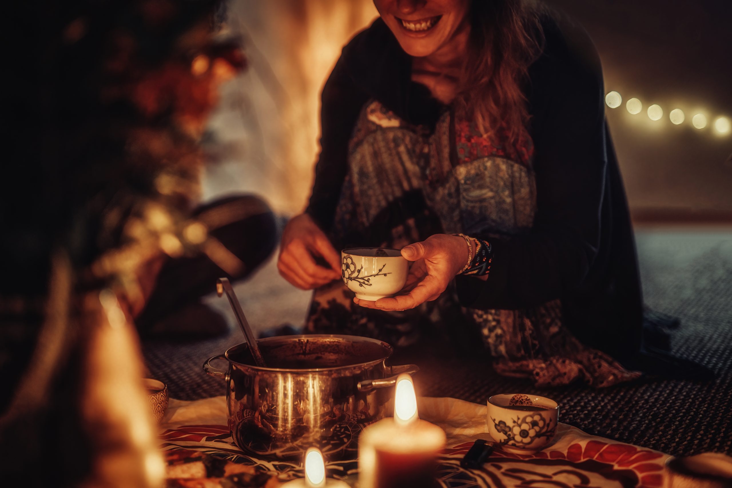 A woman smiles while serving tea from a pot, surrounded by candles and warm, ambient lighting.
