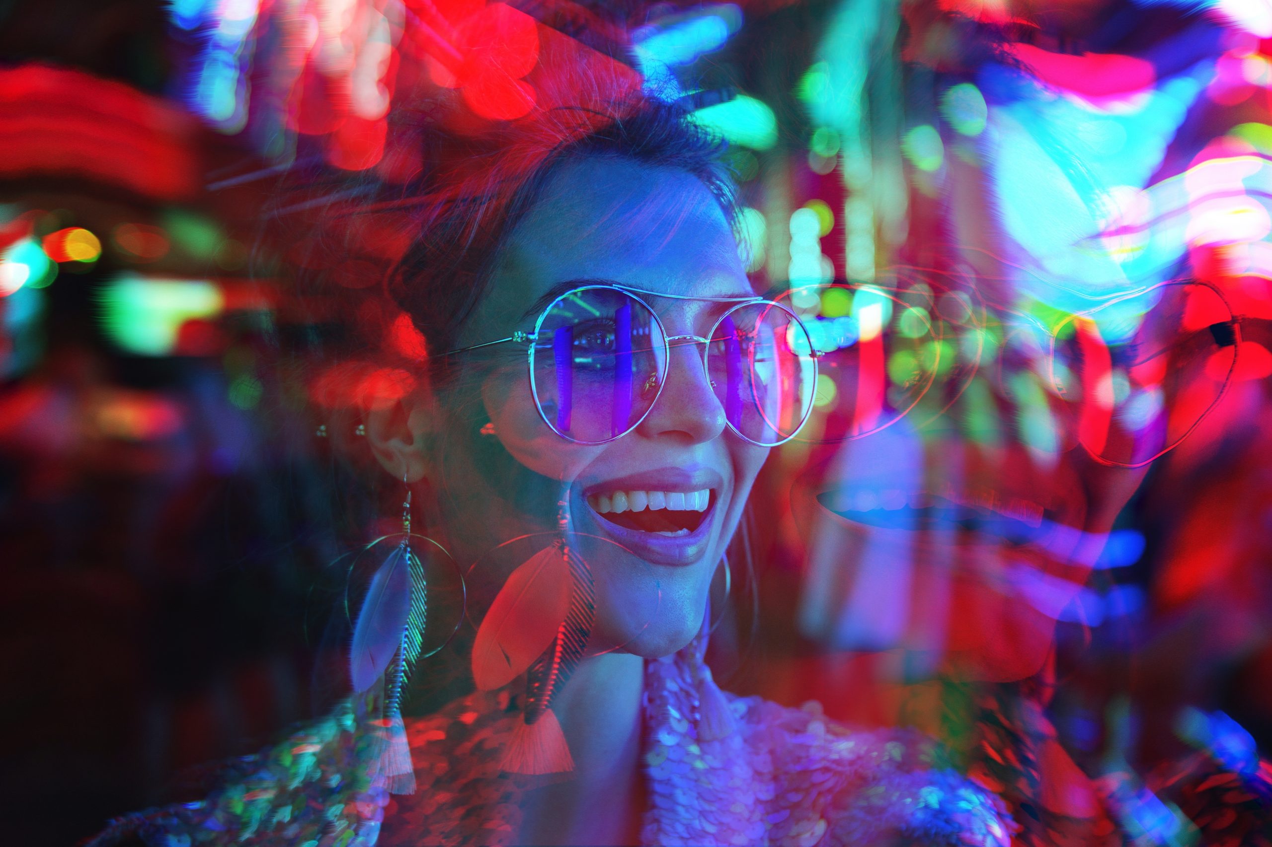 Woman with sunglasses smiles brightly amid vibrant, colorful lights and reflections.