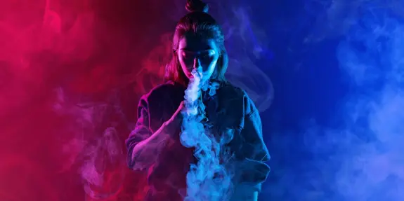 A woman with a top knot exhales colorful smoke against a vibrant red and blue background.