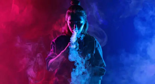 A woman with a top knot exhales colorful smoke against a vibrant red and blue background.