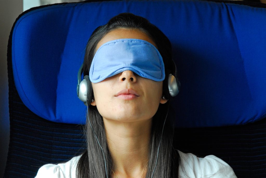 Woman wearing a sleep mask and headphones, relaxing in a blue upholstered chair.