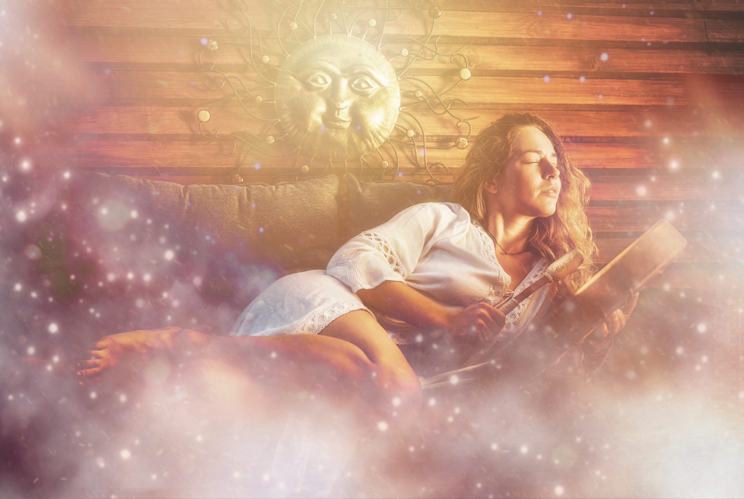 A woman in a white dress relaxes on a couch, reading, with a sun sculpture and dreamy, ethereal background.