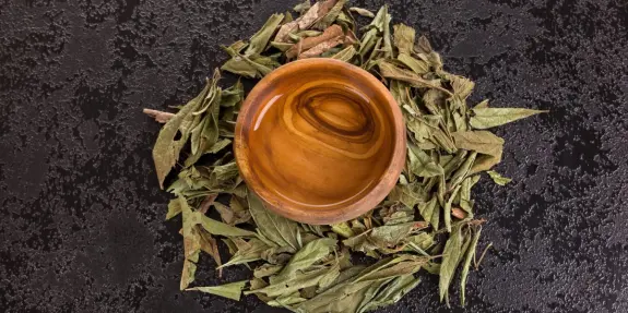 A wooden bowl filled with liquid sits atop a bed of dried leaves on a textured black surface.