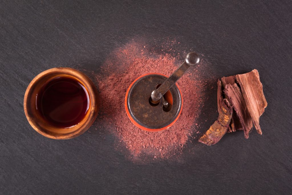 A wooden bowl with liquid, a metal container with powder, and a piece of bark on a dark surface.
