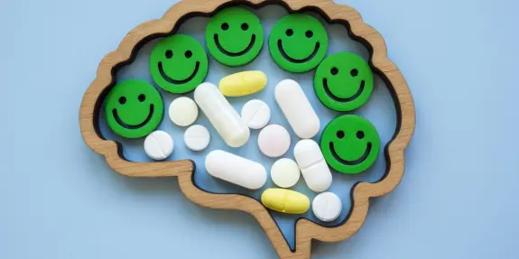 Wooden brain shape filled with colorful pills and green smiley faces on a light blue background.