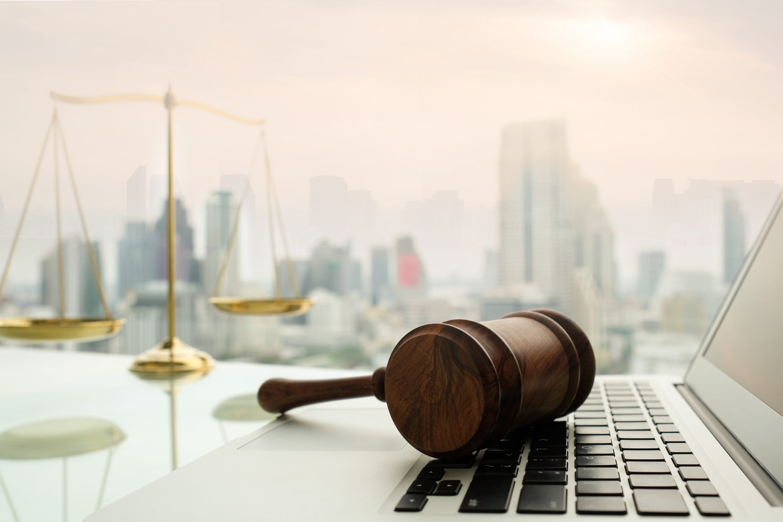 Wooden gavel rests on a laptop keyboard, with a blurred cityscape and scales of justice in the background.