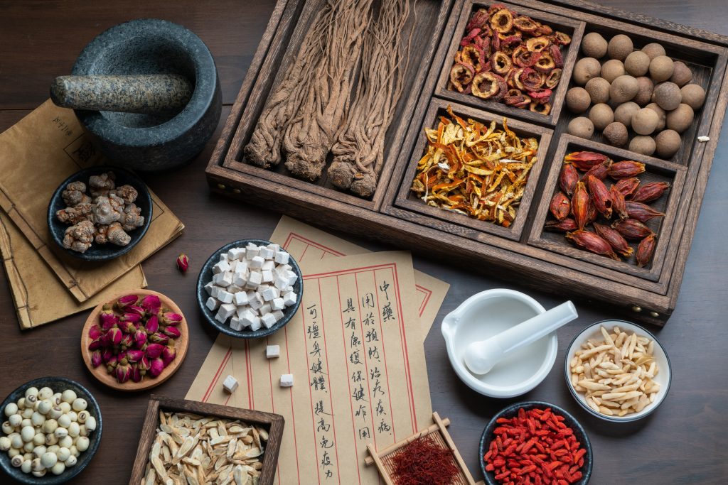 A wooden tray filled with various spices, herbs, and dried flowers, alongside a mortar and pestle and traditional papers.