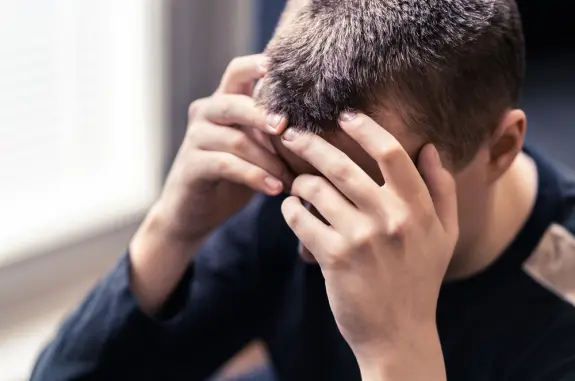 A young person sits with their head in their hands, expressing distress or frustration. Soft natural light in the background.