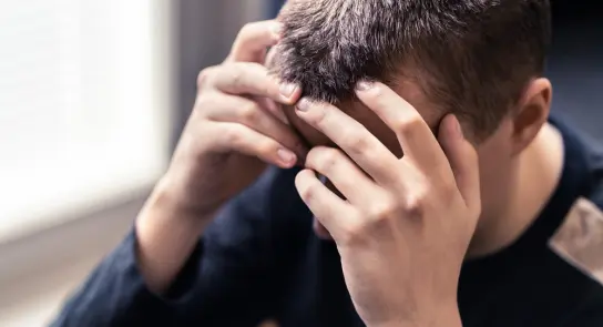 A young person sits with their head in their hands, expressing distress or frustration. Soft natural light in the background.