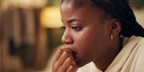 A young woman with braided hair looks pensive, biting her fingernail, in a softly lit room.