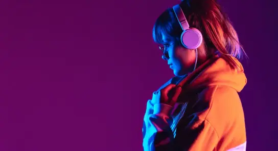 Young woman in a colorful hoodie and headphones, illuminated by vibrant purple and blue lighting.