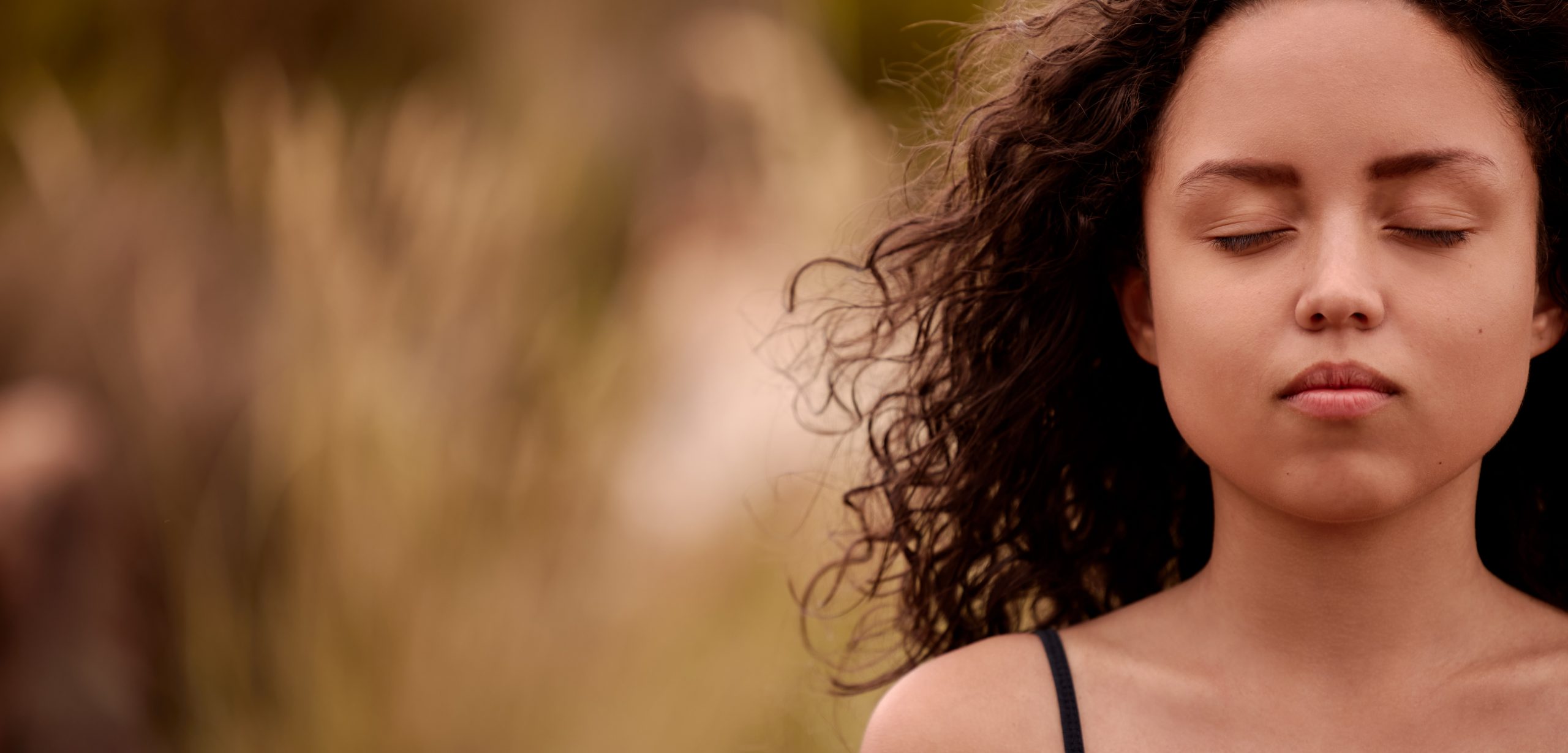 Young woman with curly hair, eyes closed, serene expression, surrounded by soft, blurred natural background.