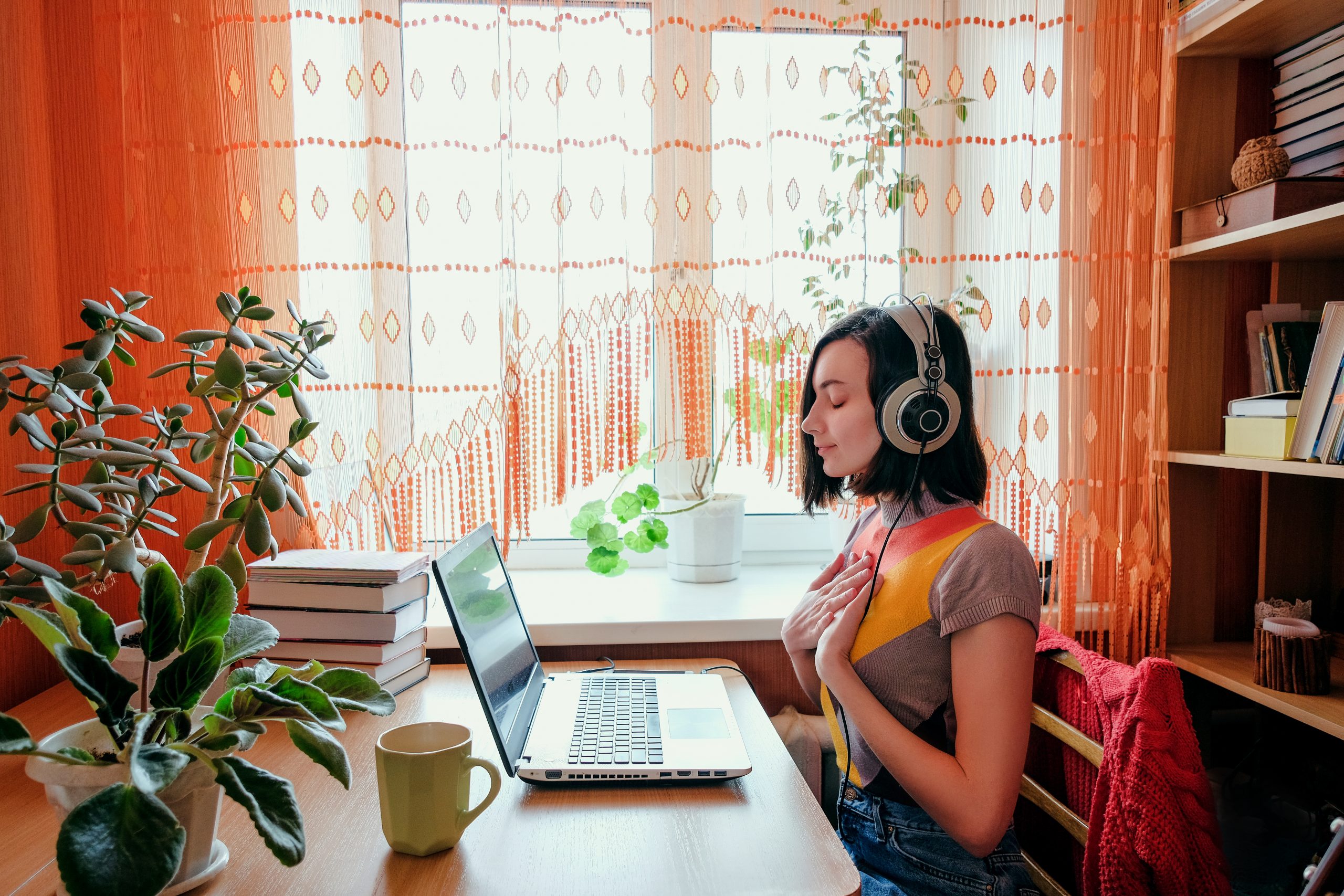 Young woman with headphones sits at a desk, focused on her laptop, surrounded by plants and warm sunlight filtering throug...