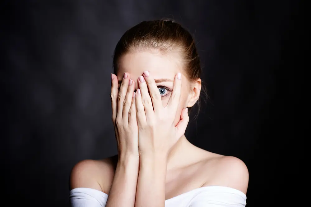 A young woman with long hair partially hides her face behind her hands, revealing a wide-eyed expression against a dark ba...