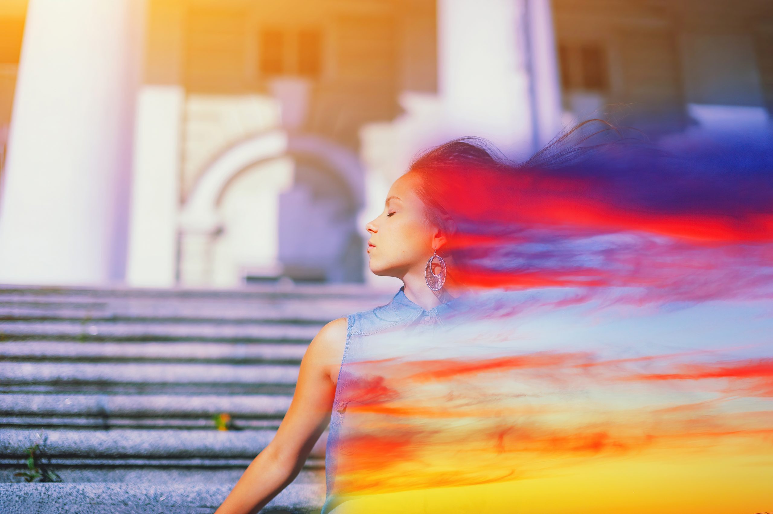A young woman stands on steps, eyes closed, with vibrant, flowing colors blending into her hair against a blurred architec...